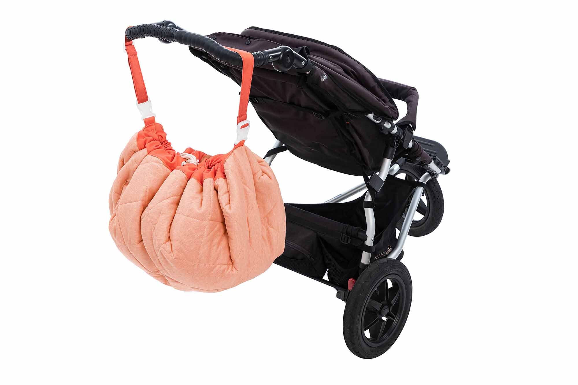 Play&Go Soft Animal Faces baby play mat, toy storage bag and diaper bag. The bag is coral with little animal faces in blue with peach noses at one side, the other side is completely coral. The diaper bag is hanging on a stroller.