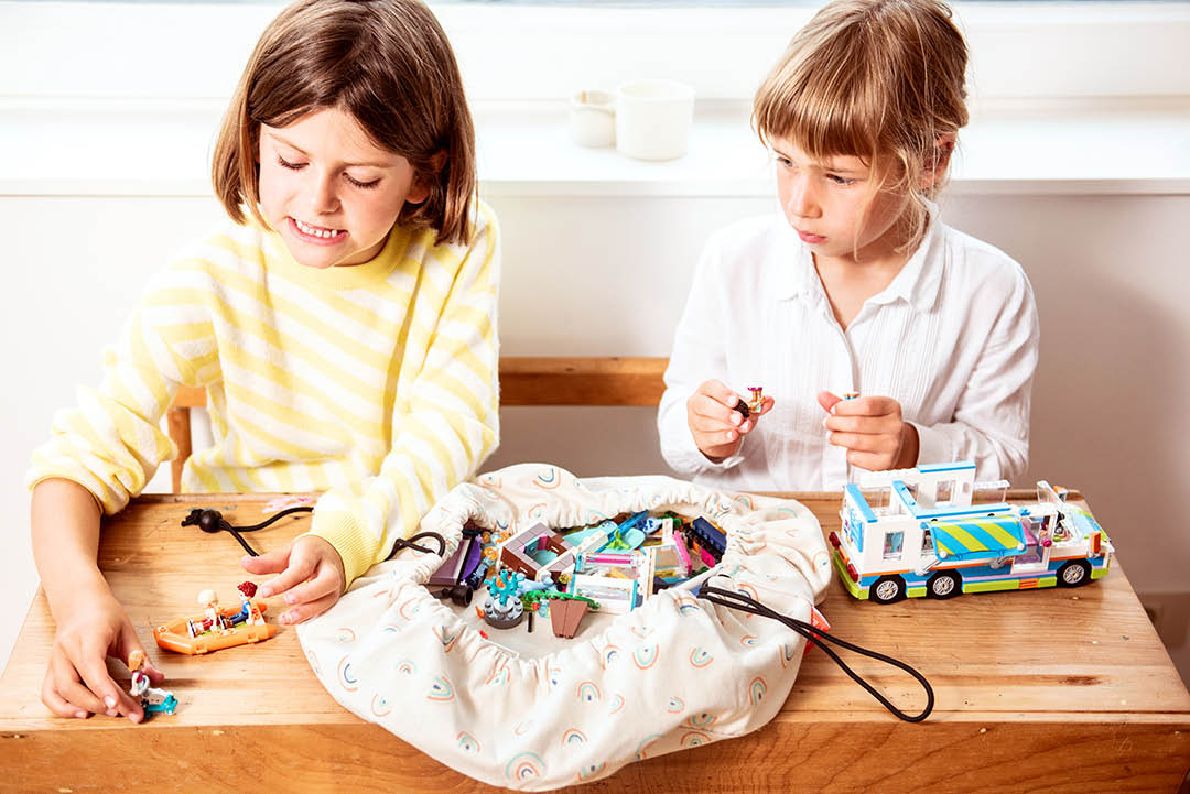 Play&Go mini storage bag, print rainbow, 2 girls playing with lego at wooden desk, bag on top of desk