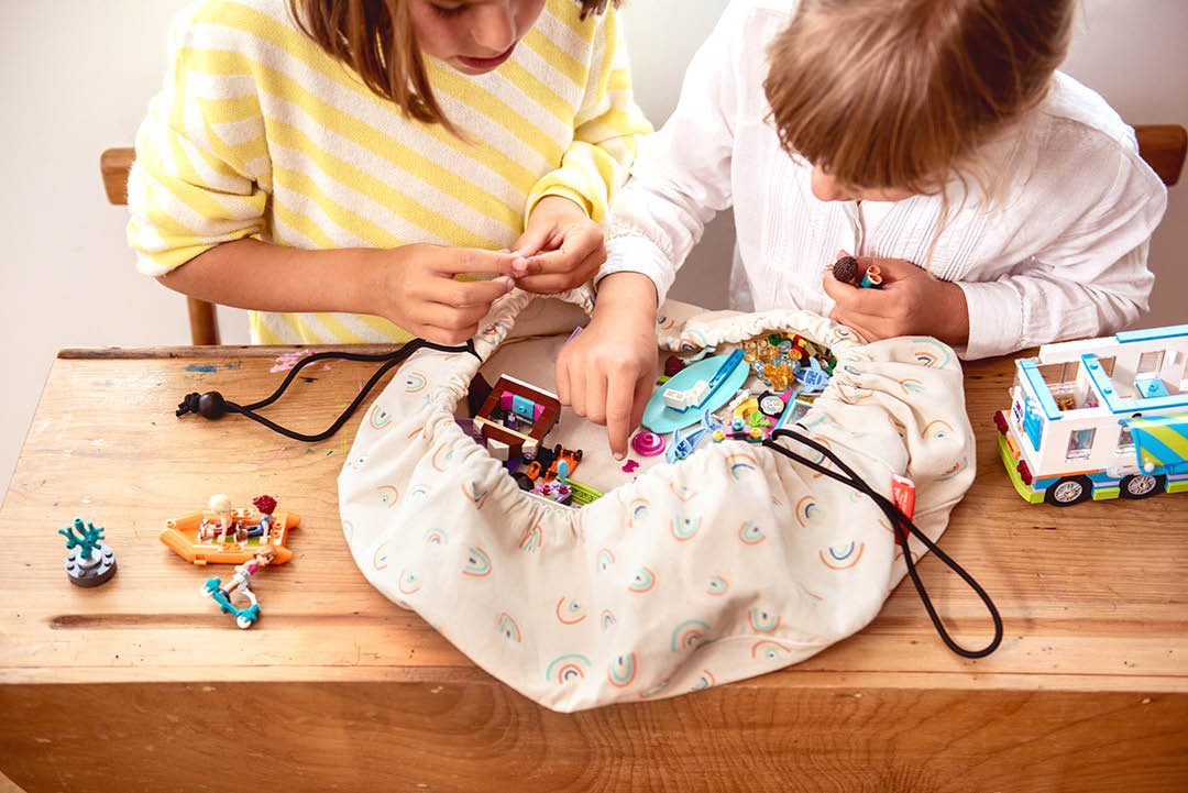Play&Go mini storage bag, print rainbow, 2 girls playing with lego at wooden desk, bag on top of desk